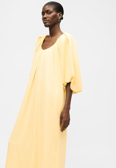 Γυναίκα που φοράει ένα χαλαρό, μακρύ παστέλ κίτρινο φόρεμα με φουσκωτά μανίκια, στέκεται μπροστά σε ένα απλό ανοιχτό γκρι φόντο.