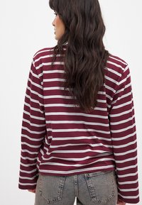 Bordeauxfarbenes Langarmshirt mit weißen horizontalen Streifen, aus weichem Stoff, mit einer lockeren Passform und rundem Halsausschnitt, getragen mit hellen Jeans.