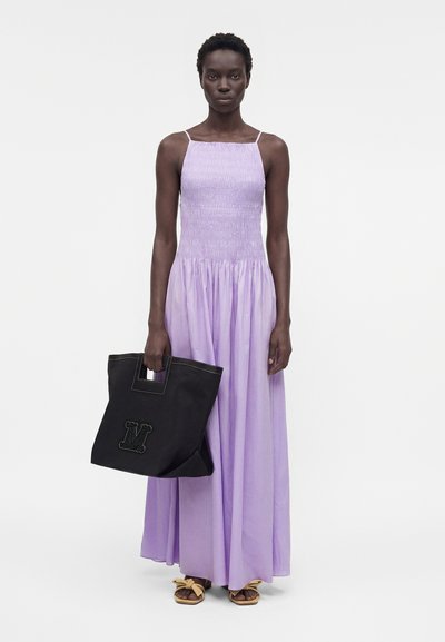 Γυναίκα που φοράει ένα μακρύ αμάνικο φόρεμα λιλά, κρατώντας μια μεγάλη μαύρη τσάντα και φοράει μπεζ σανδάλια, στέκεται μπροστά σε λευκό φόντο.