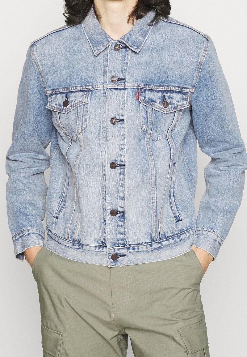 Veste en denim bleu clair présentant un devant à boutons, deux poches poitrine et un col classique. Elle a une texture légèrement usée et une coupe ajustée.