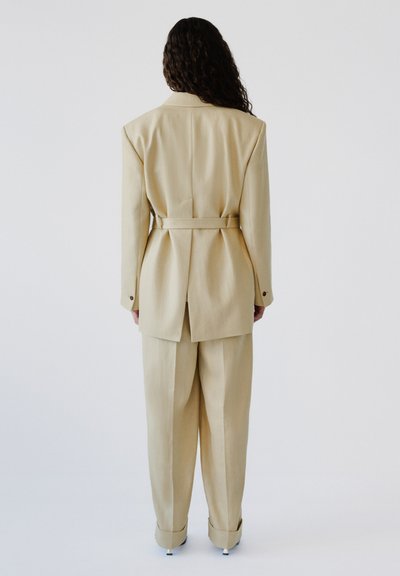 Άτομο με μακριά σγουρά μαλλιά, ορατό από την πίσω πλευρά, φορώντας μπεζ σακάκι με ζώνη και αντίστοιχο παντελόνι με φαρδιά πόδια και ρεβέρ στο τελείωμα.