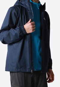 Chaqueta impermeable azul marino con capucha, puños elásticos y cremallera en la parte delantera, superpuesta sobre una capa base texturizada de color azul brillante.