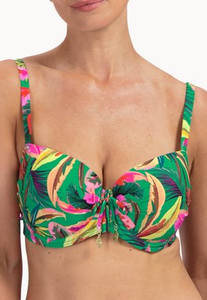 Žena má na sobě zelený horní díl bikin s barevnými tropickými listovými vzory a zlatými přívěsky ve tvaru listů uprostřed vpředu.