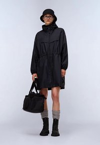 Chaqueta impermeable negra con cintura ajustable mediante cordón, mangas largas y dos bolsillos delanteros. Combinada con un sombrero tipo bucket negro y botas militares negras.