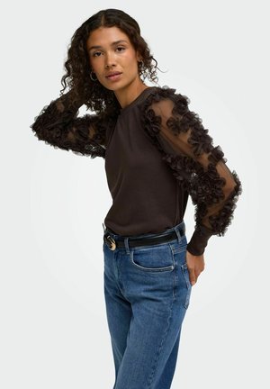 Femme aux cheveux bouclés portant un haut noir avec des manches bouffantes transparentes à motifs floraux et un jean bleu, posant avec une main dans les cheveux.