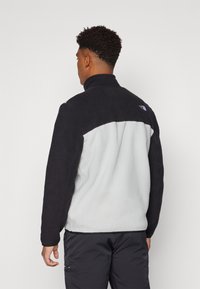 Fleecejack ontworpen met een zwart bovenstuk en een lichtgrijs onderstuk, met een hoge kraag en het logo van The North Face op de schouder.