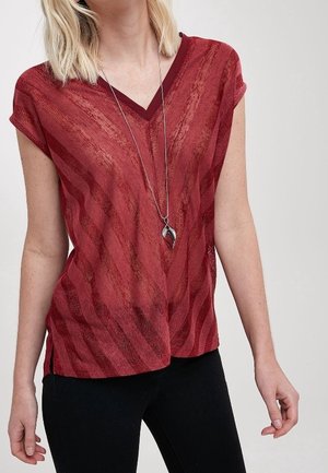Femme portant un t-shirt rouge à manches courtes, à motif et col en V, avec un collier avec pendentif en argent, associé à un pantalon noir.