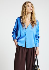 Woman with wavy blonde hair wearing a blue satin button-up shirt and brown wide-leg pants, holding a black handbag in her left hand.