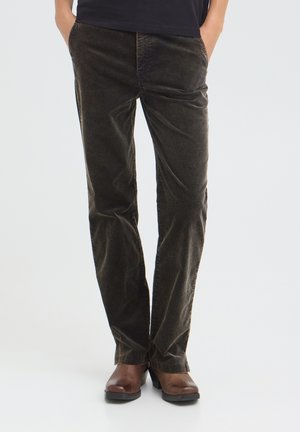 Personne portant un pantalon en velours côtelé marron foncé et des bottes en cuir marron, debout les mains dans les poches sur un fond uni.