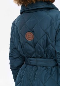 Teal gestepte jas met een ceintuur in de taille, met een hoge kraag en een cirkelvormige bruine leren patch met de tekst "blue shadow."