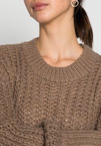 Brązowy sweter dzianinowy z teksturowanym, grubym splotem, szerokim, prążkowanym wykończeniem dekoltu oraz luźnym krojem. Udekorowany widocznymi szwami i miękkimi włóknami.