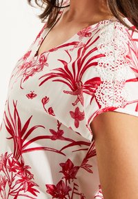 Hvid bluse med rødt blomsterprint, V-udskæring og blonde detaljer på ærmerne. Glat tekstur med botaniske detaljer og elegant design.
