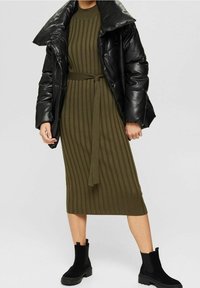 Svart blänkande pufferjacka, olivgrön ribbad stickad klänning med bälte, knälång. Svarta ankelskor med texturerad sula.
