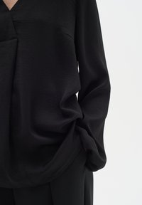 Maglietta nera a maniche lunghe con scollo a V, vestibilità ampia e texture morbida, con drappeggio delicato e dettaglio della tasca laterale.