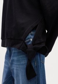 Sweat-shirt noir avec une fente latérale nouée en nœud sur le détail de la poche d’un jean en denim bleu.