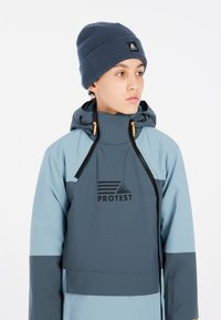 Veste imperméable bleu clair et gris foncé avec capuche, arborant un logo noir "PROTEST" et des tirettes de fermeture éclair orange. Portée avec un bonnet bleu foncé.