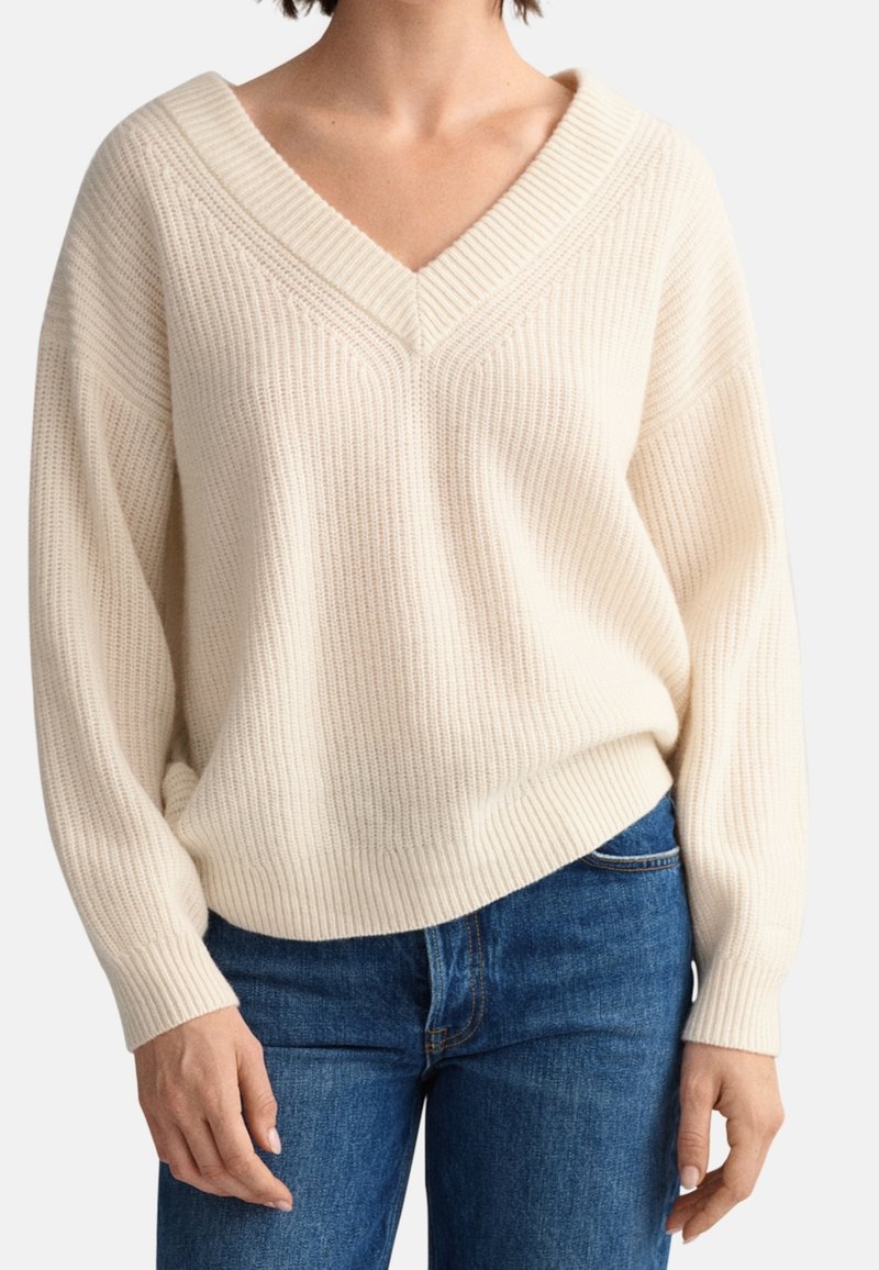 Hellbeige Strickpullover mit V-Ausschnitt und gerippter Textur; lockere Passform mit langen Ärmeln, kombiniert mit blauen Jeans.