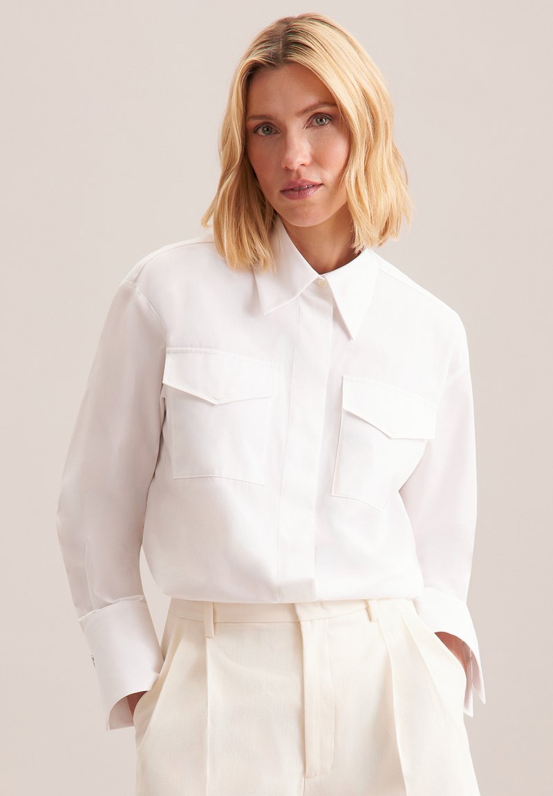 Camicia bianca a maniche lunghe con colletto, due tasche frontali e polsini abbottonati, abbinata a pantaloni a vita alta color crema. Tessuto morbido.