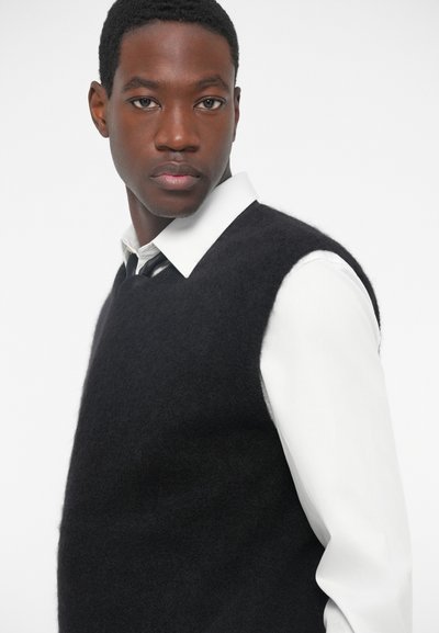 Črn brezrokavnik iz mehkega, strukturiranega blaga, nošen preko bele srajce s kragnico, viden s strani.