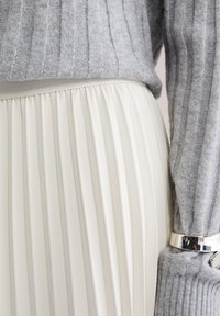 Tuva sudraba aproce uz pelēkas ribotas džempera piedurknes tuvplānā un baltas kroku svārku jostasvietas detalizēts skats.