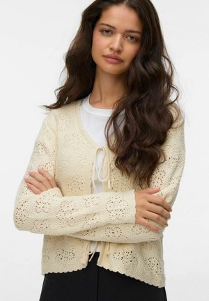 Ung kvinde med langt bølget mørkt hår iført en cremefarvet strikket cardigan over en hvid top, med armene krydset, foran en ensartet lys baggrund.