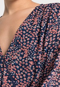 Abito floreale con una base blu navy e fiori rosa e bianchi. Presenta una profonda scollatura a V e un tessuto morbido e testurizzato con maniche drappeggiate.