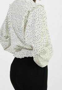 Witte blouse met zwarte stippen, gerimpelde kraag, elastische taille en volumineuze mouwen. Zwarte broek zichtbaar aan de onderkant.