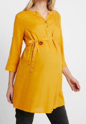 Gravid kvinde iført en sennepsgul graviditetstunika med trekvartlange ærmer, knappet halsudskæring og bundet bælte i taljen over sorte bukser.