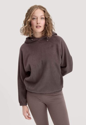Junge Frau mit lockigem Haar, die einen braunen Fleece-Hoodie und passende Leggings trägt, steht vor einem einfarbigen hellen Hintergrund.