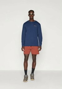 Tengerészeti hosszú ujjú póló logóval, rozsdabarna rövidnadrág, fekete bokazokni és barna sportcipő. Modell napszemüveget visel. Egyszerű háttér.