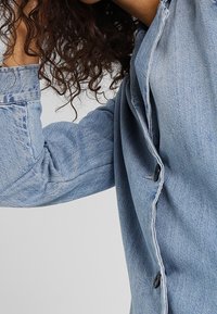 Camisa de denim azul claro con un cierre de botones en la parte delantera, mangas largas con detalles en los puños y textura sutil. Los botones son negros para crear contraste.