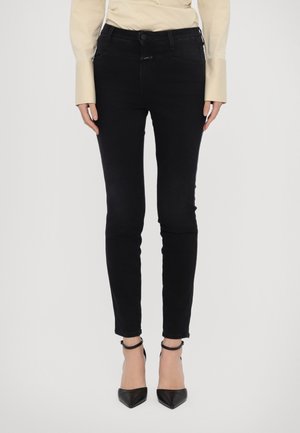 Mujer vistiendo pantalones negros ajustados, camisa beige de manga larga y tacones negros con correa en el tobillo y punta afilada, posando contra un fondo claro.