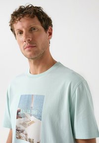 Camiseta de algodón de color azul claro con una impresión fotográfica de una silla junto al mar y el texto "SALSA JEANS". Diseño de manga corta y cuello redondo.