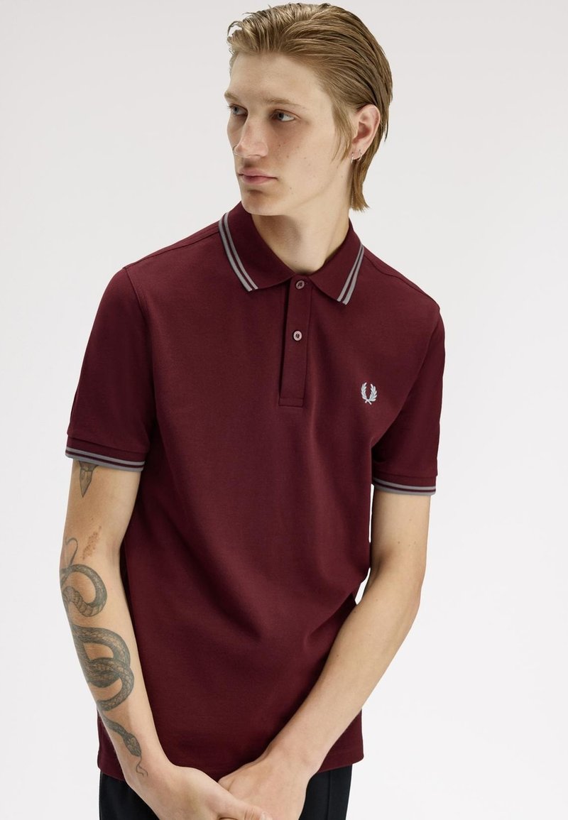 Hombre joven con cabello claro que lleva una camisa polo granate con cuello y puños de manga a rayas grises, mostrando un tatuaje de serpiente en el antebrazo izquierdo.