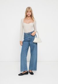 Junge Frau mit blonden Haaren, die ein beiges Langarmoberteil, hochgeschnittene, weit geschnittene blaue Jeans, schwarze Ballerinas trägt und eine weiße Handtasche am Arm hält.