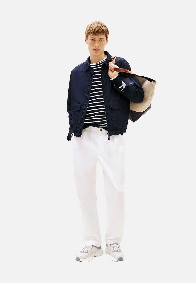 Chaqueta encerada azul marino sobre una camiseta de rayas negras y blancas, pantalones blancos y zapatillas grises; el modelo sostiene un bolso de tote beige y azul marino.