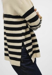 Maglione lavorato a maglia con righe orizzontali nere e crema, dotato di una fessura laterale per facilitare i movimenti, abbinato a jeans di denim scuri.