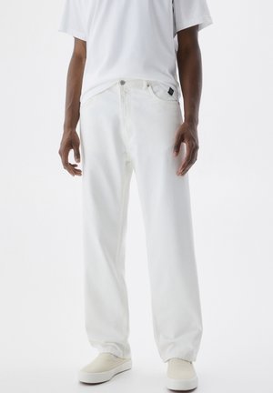 White denim jeans with a relaxed fit and visible stitching. Features side pockets and a classic waistband. Paired with beige slip-on shoes.