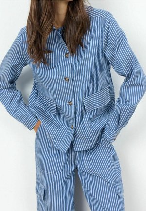 Femme portant une veste boutonnée à rayures verticales bleues et blanches et un pantalon assorti, les mains dans les poches, devant un fond uni.