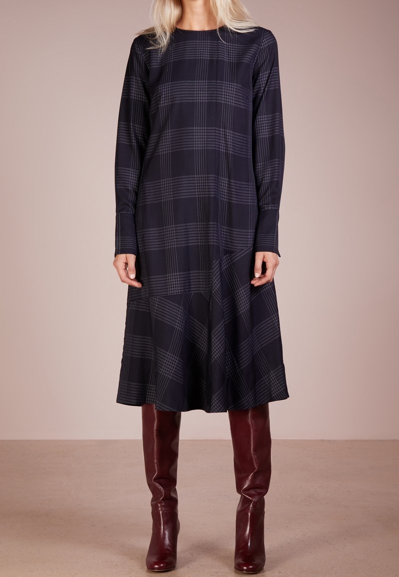 Femme portant une robe midi à manches longues à carreaux noirs associée à des bottes en cuir marron foncé montant jusqu'aux genoux sur un fond neutre.