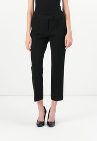 Zwarte op maat gemaakte broek met een aansluitende tailleband, plooidetails en een cropped lengte, gecombineerd met zwarte laarzen van gepatineerd leer met hoge hakken.