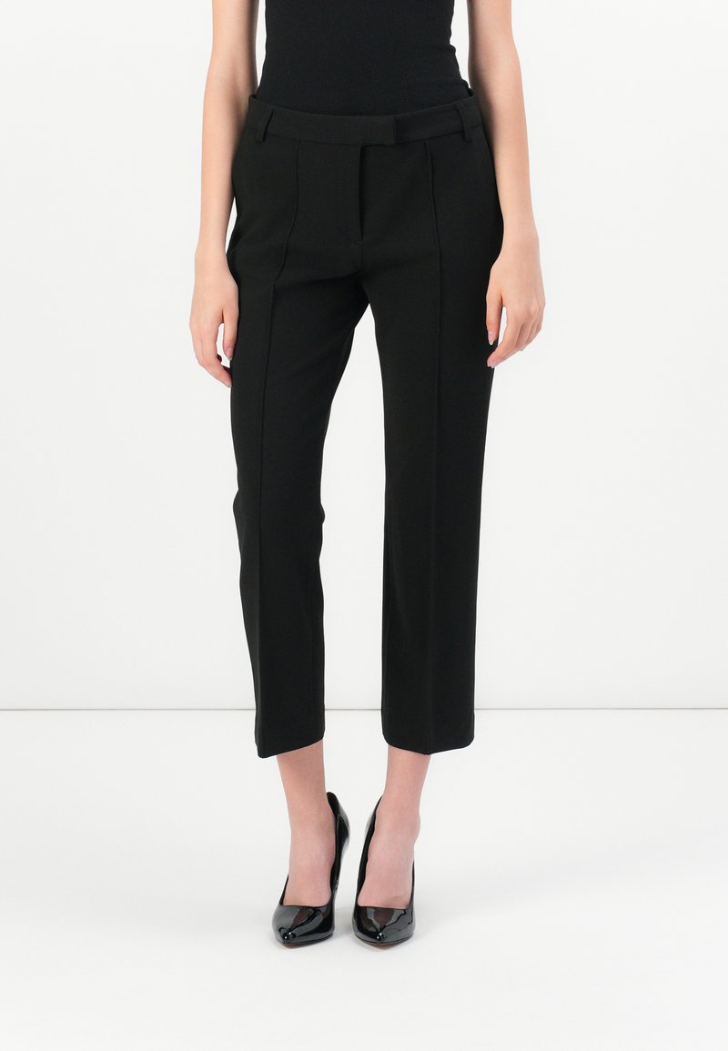 Zwarte op maat gemaakte broek met een aansluitende tailleband, plooidetails en een cropped lengte, gecombineerd met zwarte laarzen van gepatineerd leer met hoge hakken.