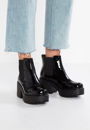 Glanzende zwarte plateau enkellaarsjes met dikke hakken, gedragen met cropped lichtblauwe jeans met rafelrand op een witte vloer.