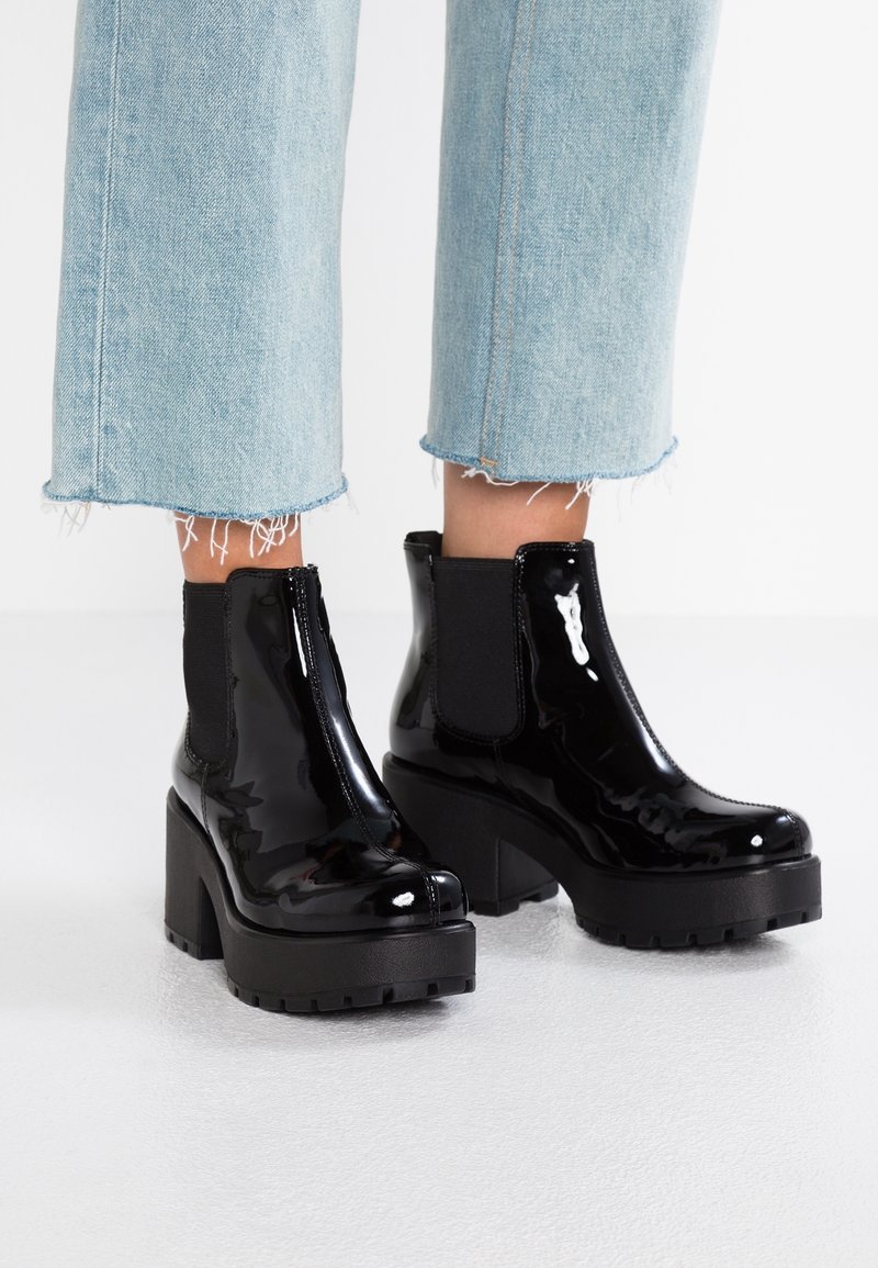 Bottes noires à plateforme brillantes avec talons épais portées avec un jean court bleu clair à ourlet effiloché sur un sol blanc.