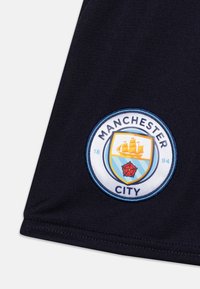 Grb nogometnega kluba Manchester City, okrogla zasnova z modro in rumeno grafiko ladje, rdečo vrtnico ter besedilom na temnem tekstilnem ozadju.
