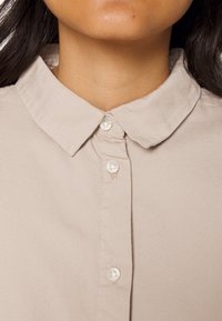 Chemise col historique beige clair avec une fermeture à boutons sur le devant, ayant une texture lisse et de petits boutons ronds.