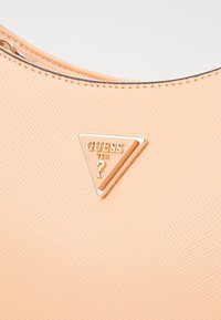 Torbica breskvaste boje s teksturiranom površinom, koja sadrži trokutasti zlatni metalni logo s natpisom "GUESS USA."