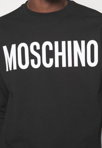 Sweat-shirt noir avec le grand texte blanc "MOSCHINO" sur le devant. Confectionné en matériau doux avec un col rond et des manches longues.