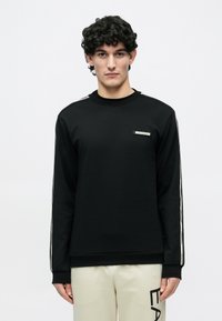 Juodas ilgomis rankovėmis džemperis apvaliu kaklu, su šoninėmis juostelėmis ir mažu logotipo pleistru ant krūtinės. Minkštas audinio tekstūra.