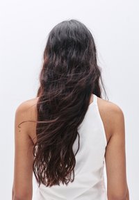 Lang, golvend donker haar valt naar beneden over de rug van een persoon die een witte mouwloze top draagt tegen een effen, lichte achtergrond.